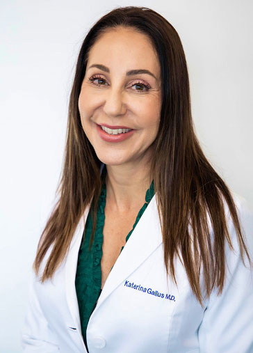 Dr. Katerina Gallus smiling in white coat. She offers a range of procedures, including breast augmentation breast reduction and much more. Options include both saline and silicone implants.