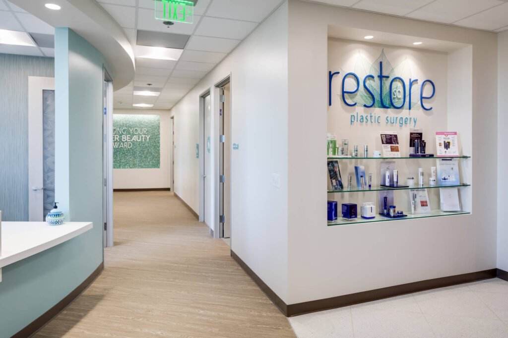 Well-lit front desk area at plastic surgery office with pale blue and white walls, a display of skincare products, and the practice name "Restore SD Plastic Surgery" on the wall.