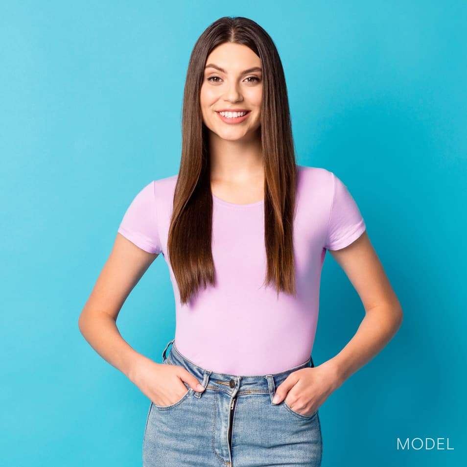 Image of model standing against turquoise background with hands in pockets smiling where a light purple t-shirt and jeans.
